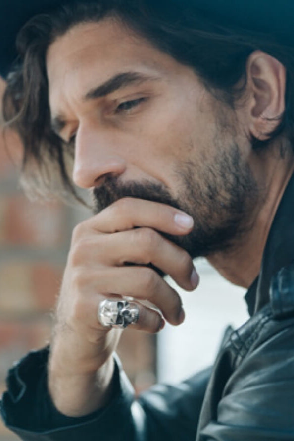 a man sits with his hand on his chin. He has thick dark  hair. He is thinking. On his hand he is wearing a large skull ring on his little finger.